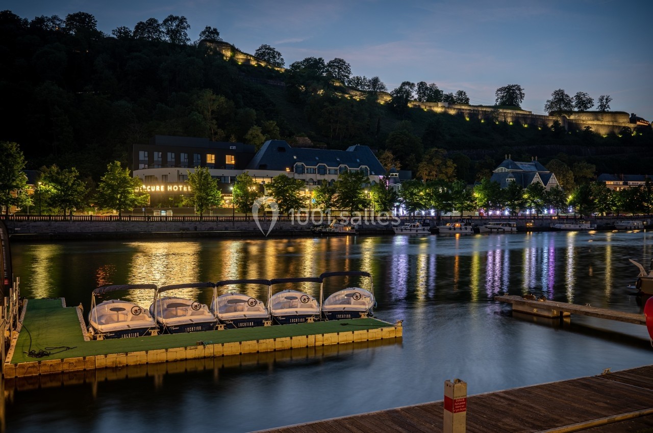 Bateaux électriques amarrés sur une rivière au crépuscule, avec des bâtiments éclairés et une colline en arrière-plan.