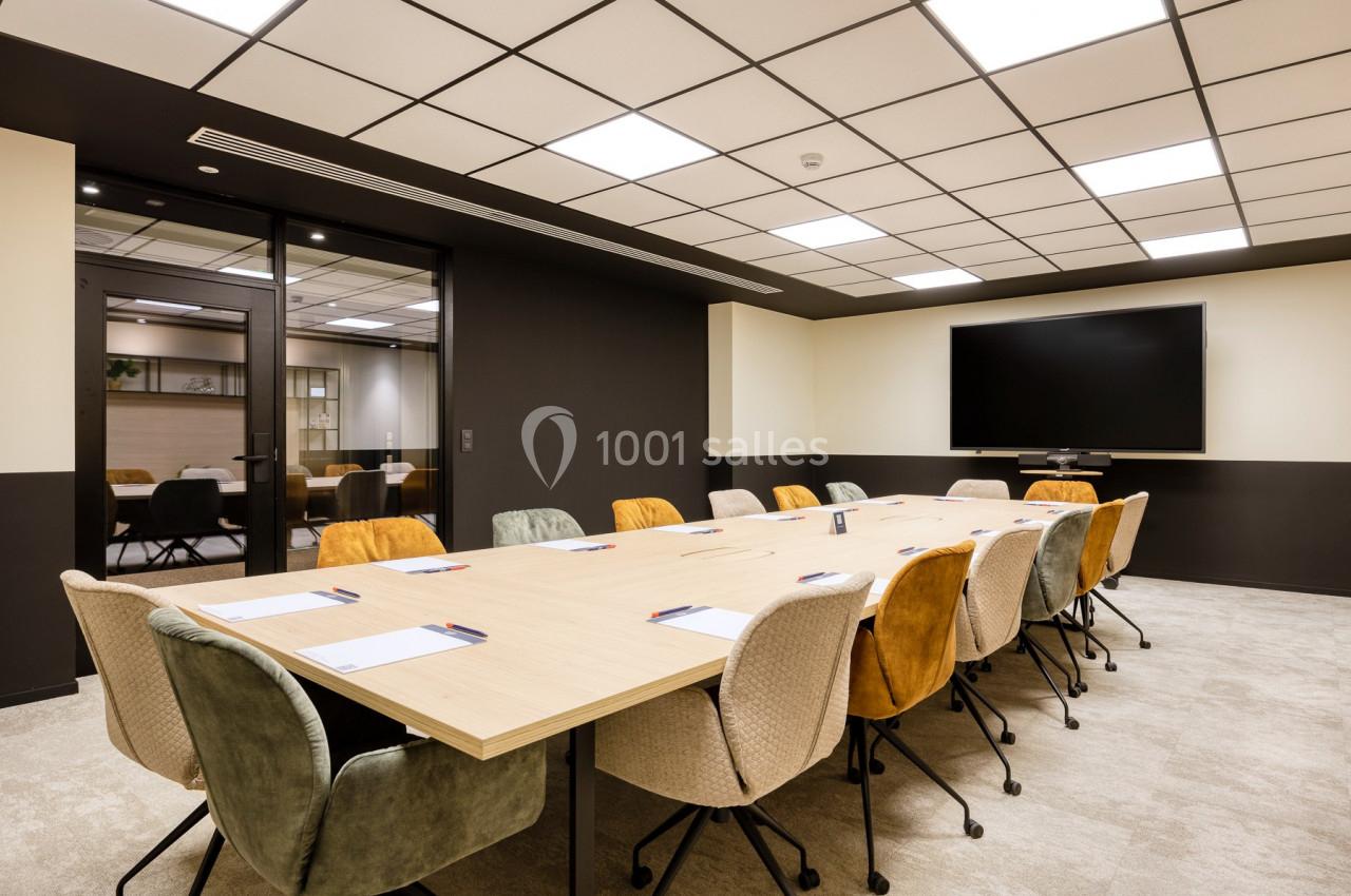 Salle de réunion moderne avec une grande table rectangulaire entourée de chaises colorées et un écran mural.