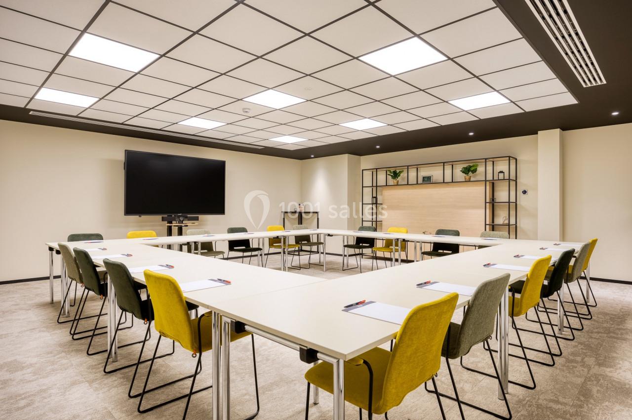 Salle de réunion lumineuse avec tables disposées en U, chaises colorées, écran mural et étagères en arrière-plan.