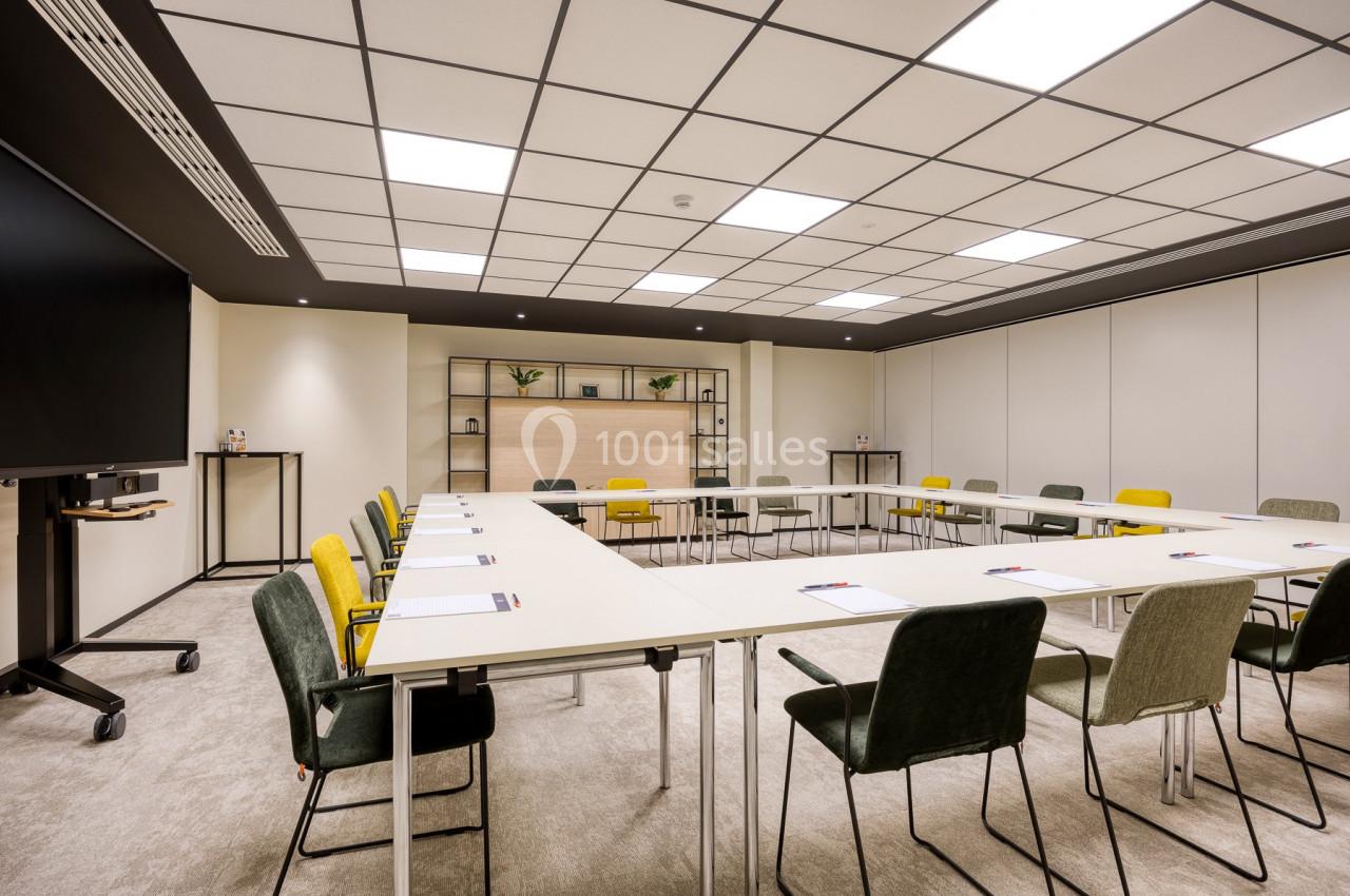 Salle de réunion lumineuse avec tables disposées en U, chaises colorées et étagères décoratives en arrière-plan.