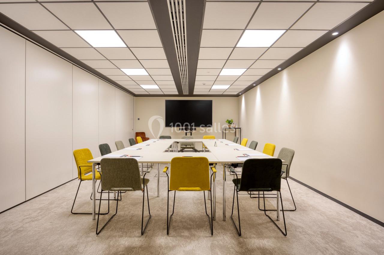 Salle de réunion moderne avec table rectangulaire, chaises colorées, écran mural et éclairage au plafond.