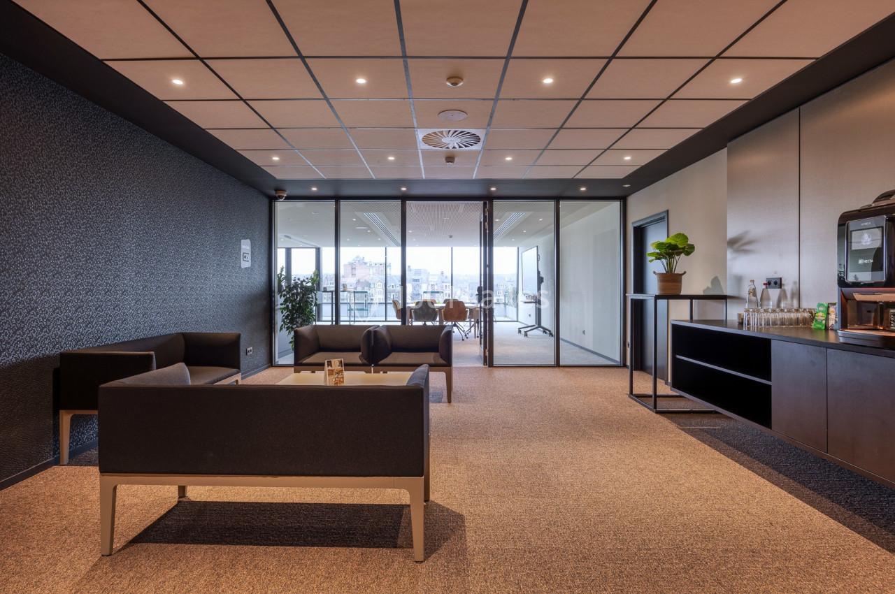 Salle d'attente moderne avec canapés, table basse, plantes et vue sur une salle de réunion vitrée.