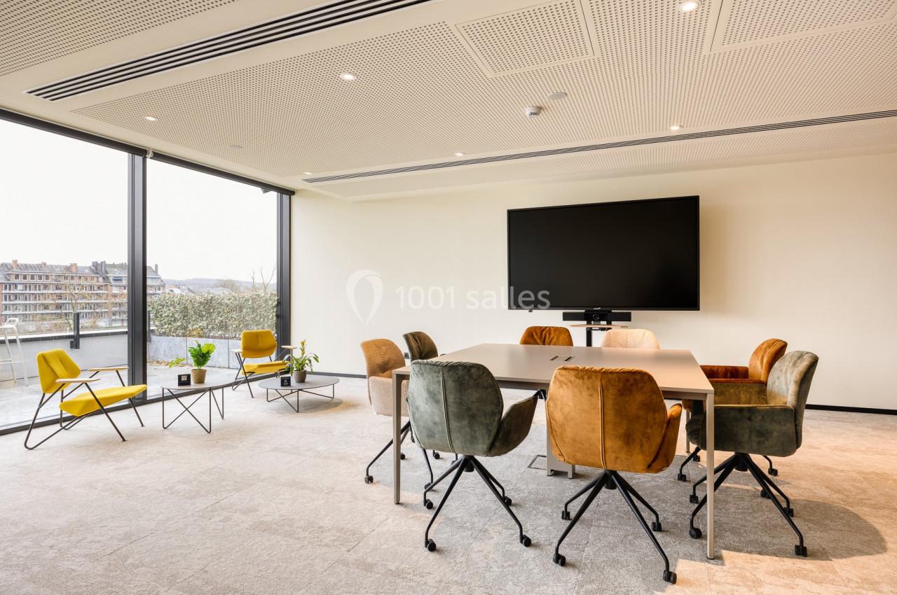 Salle de réunion lumineuse avec table, chaises colorées, écran mural et grandes baies vitrées donnant sur l'extérieur.
