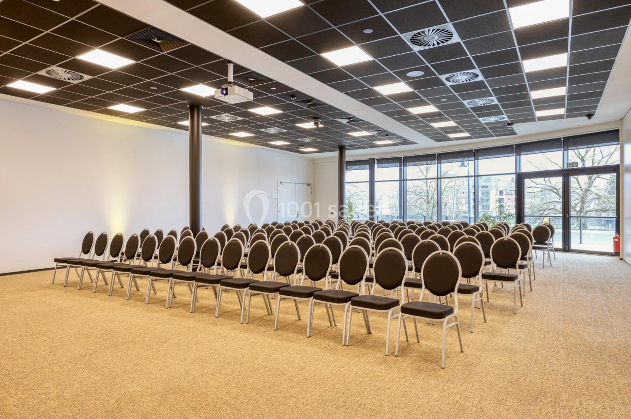 Salle de conférence lumineuse avec des rangées de chaises noires alignées face à un écran de projection.