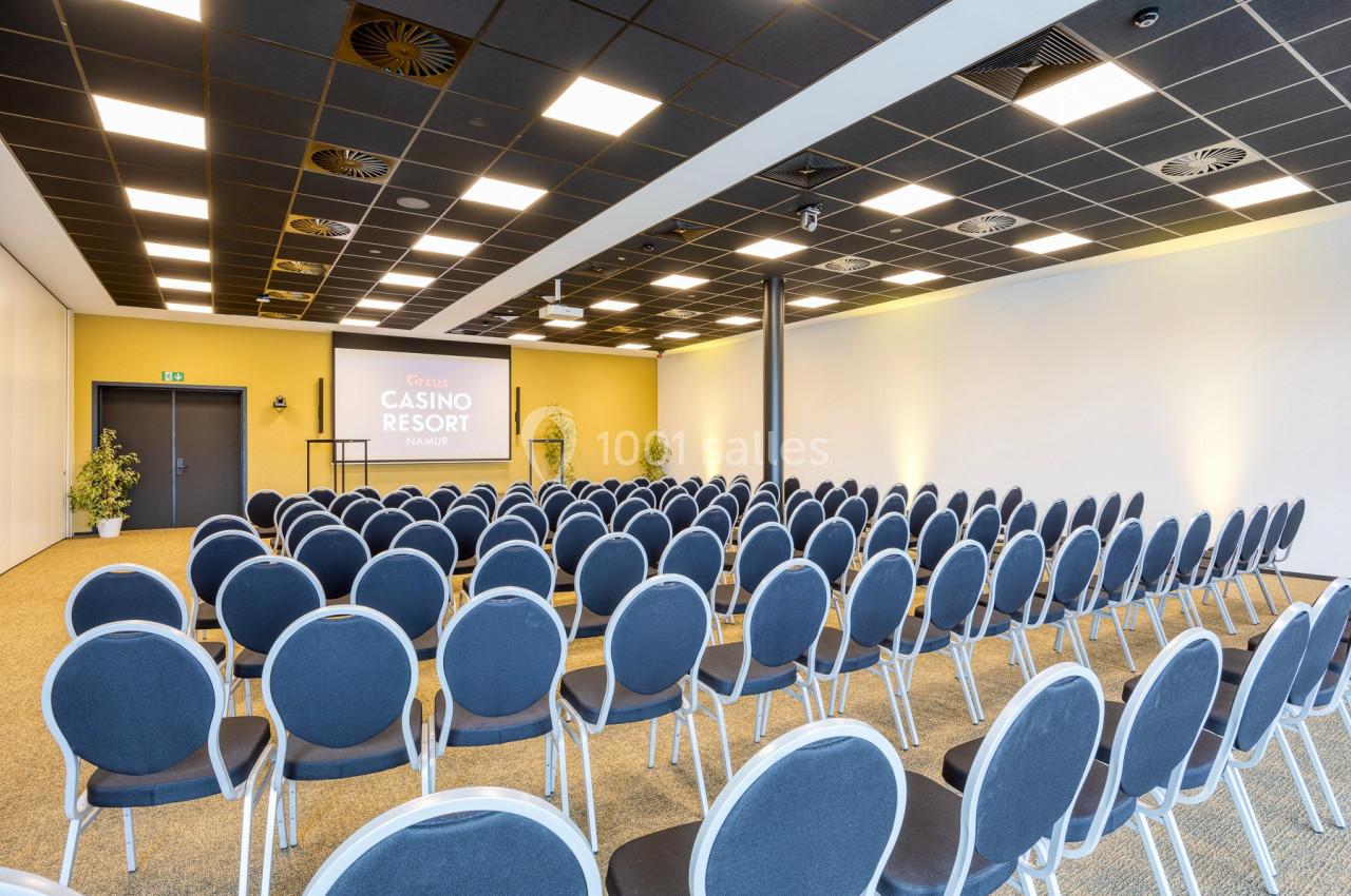 Salle de conférence avec rangées de chaises bleues alignées face à un écran de projection et un pupitre.