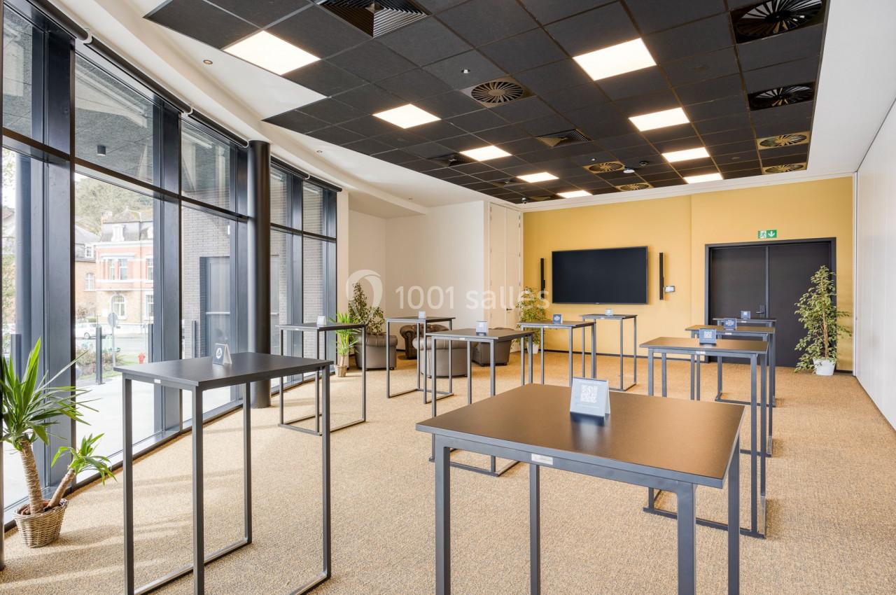 Salle lumineuse avec tables hautes espacées, grandes baies vitrées, plantes et écran mural.