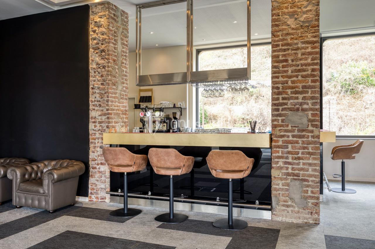 Bar avec comptoir en bois, tabourets en cuir, murs en briques apparentes et grandes fenêtres donnant sur l'extérieur.