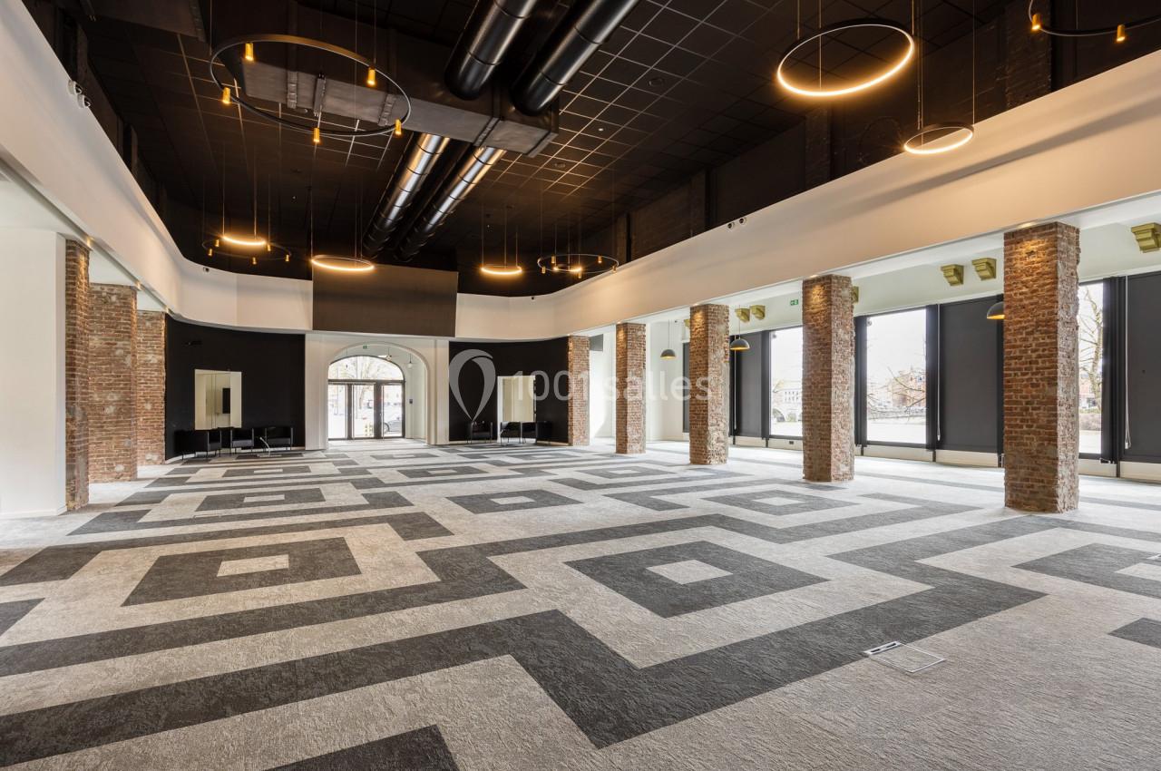 Salle spacieuse avec sol à motifs géométriques, colonnes en briques, grandes fenêtres et luminaires suspendus.