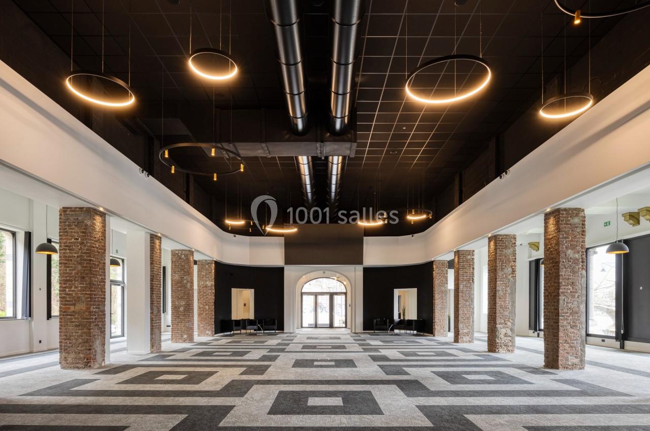 Salle spacieuse avec colonnes en briques, plafond noir, luminaires circulaires suspendus et sol à motifs géométriques.