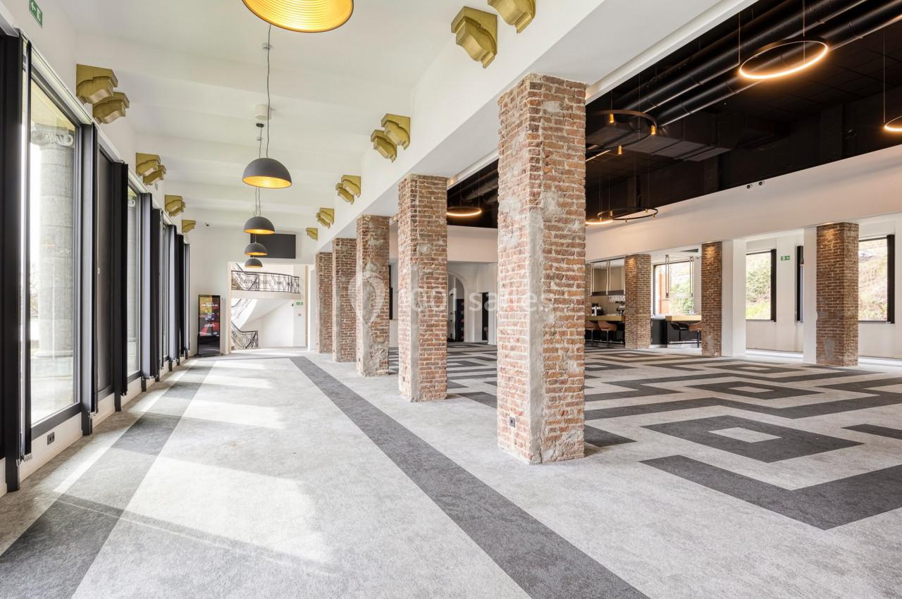 Salle lumineuse avec colonnes en briques, grandes baies vitrées, sol en motifs géométriques et plafonniers suspendus.