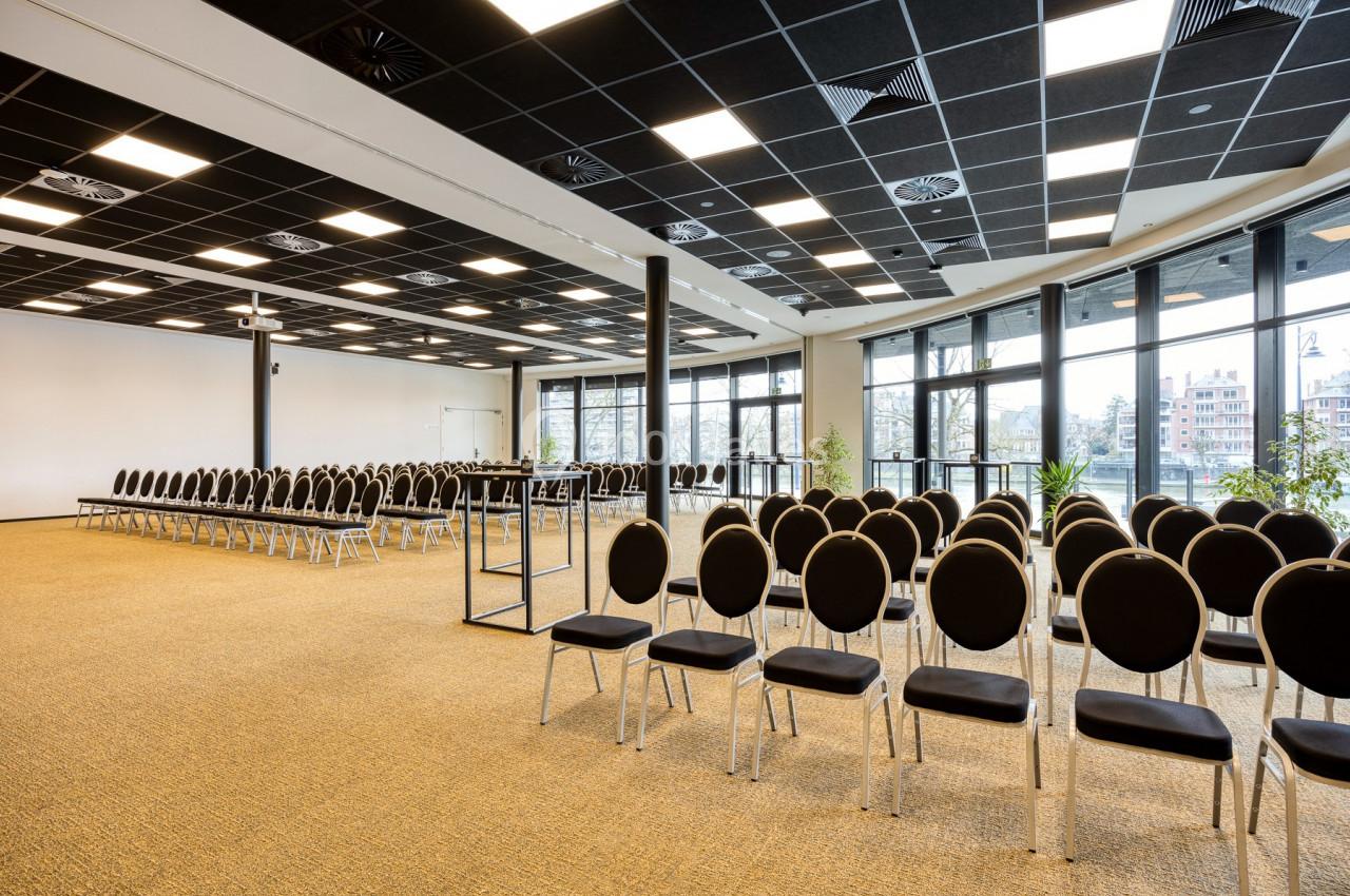 Salle de conférence lumineuse avec rangées de chaises noires alignées, grandes fenêtres et moquette beige.