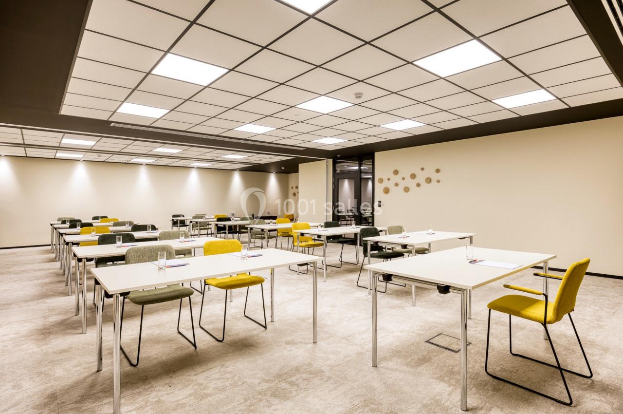 Salle de réunion lumineuse avec des tables blanches alignées, chaises colorées et un plafond à dalles.