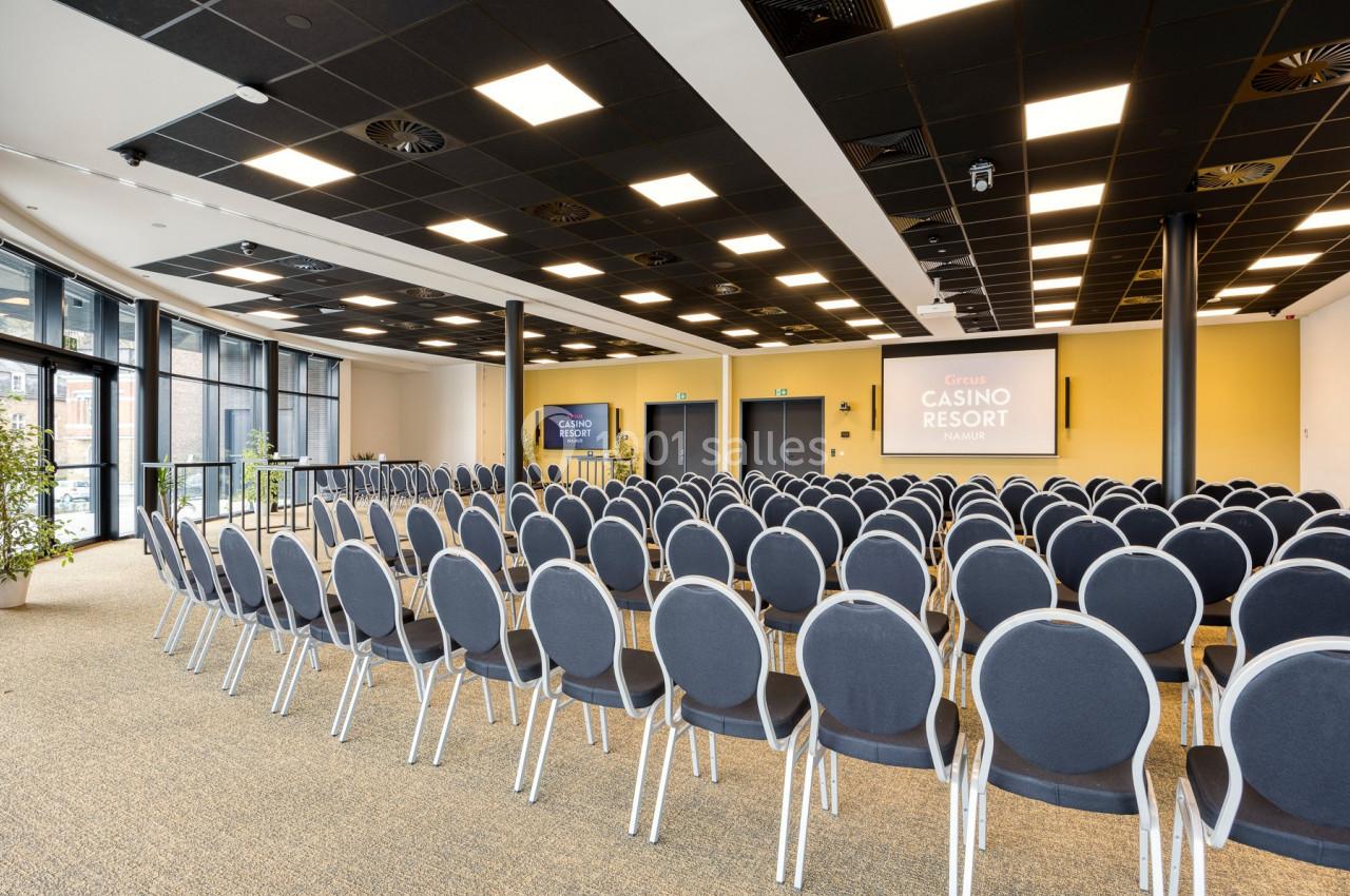Salle de conférence lumineuse avec des rangées de chaises grises alignées face à deux écrans de présentation.