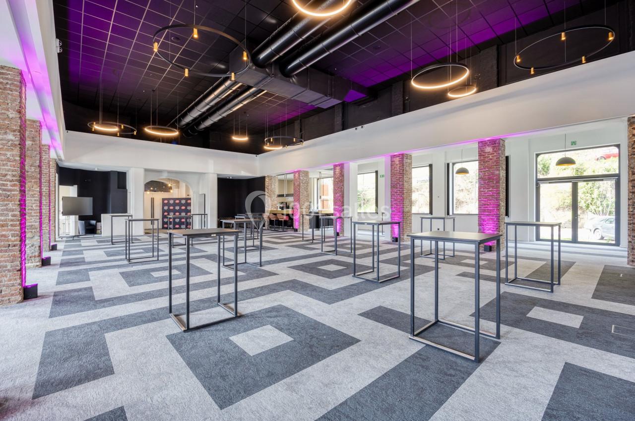 Salle moderne avec un sol à motifs géométriques, des tables hautes métalliques et un éclairage suspendu circulaire.