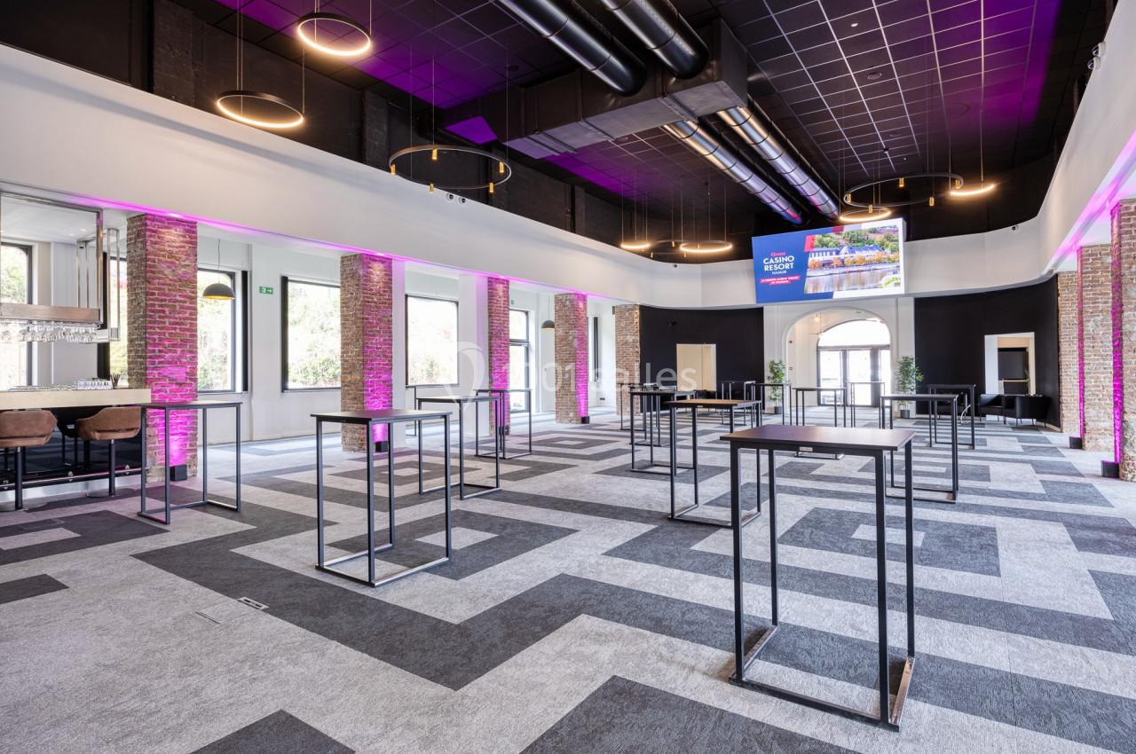Salle moderne avec tables hautes, éclairage suspendu, murs en briques et néons roses, sol à motifs géométriques.