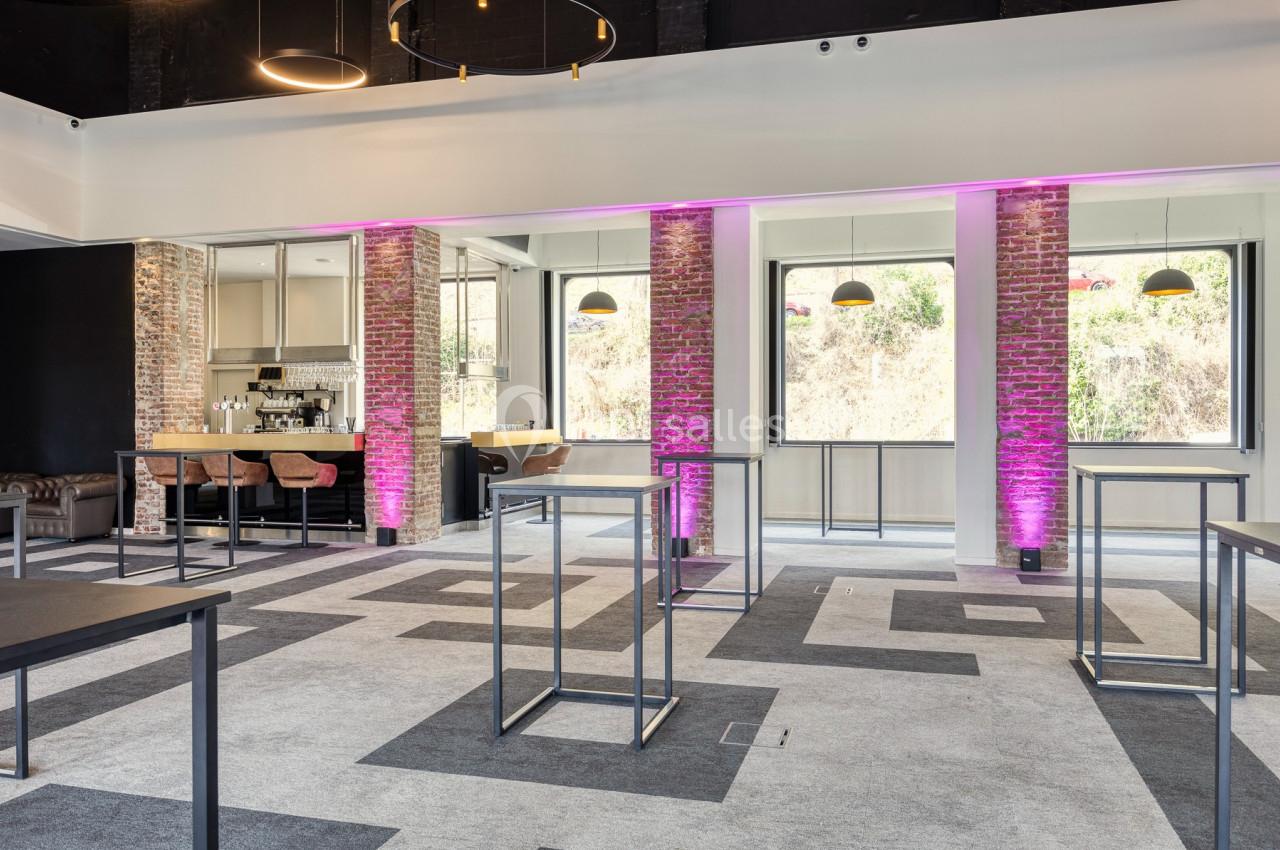 Salle lumineuse avec murs en briques, bar équipé, tables hautes et éclairage violet décoratif.