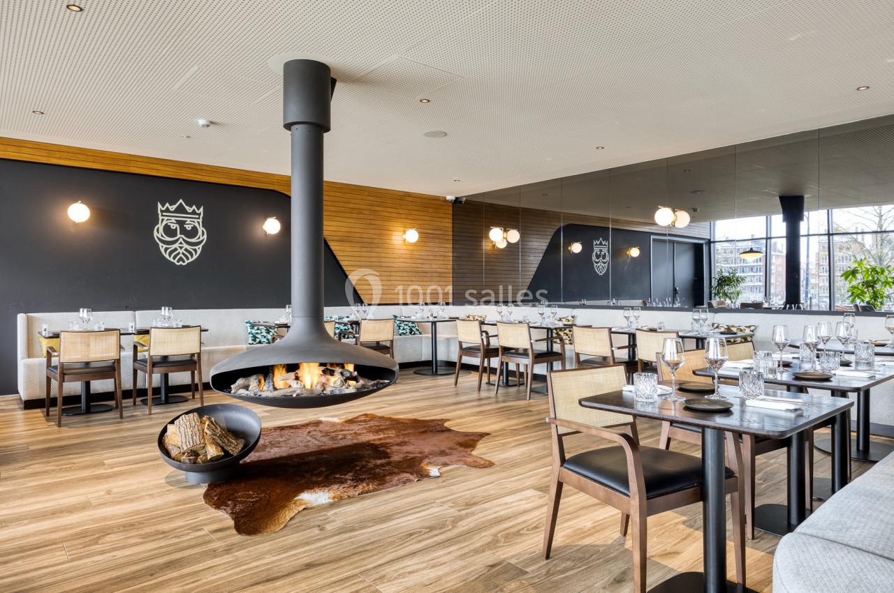Salle de restaurant moderne avec cheminée suspendue, tables dressées et décoration élégante sur fond de bois et miroirs.