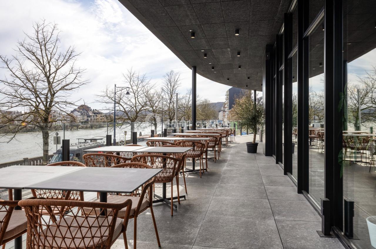 Terrasse d'un restaurant avec tables et chaises en osier, offrant une vue sur une rivière bordée d'arbres sans feuilles.