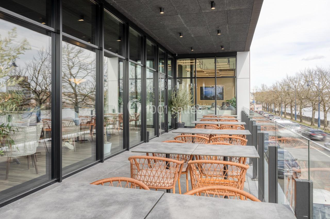 Terrasse moderne avec tables et chaises en rotin, bordée de baies vitrées et vue sur une rue arborée.