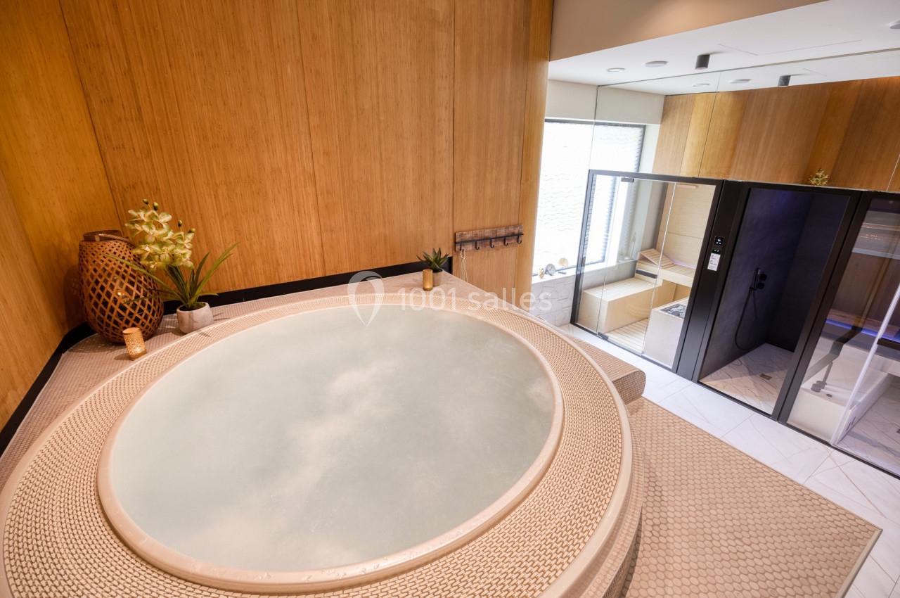 Bain à remous dans une pièce en bois clair avec une douche et un sauna vitré en arrière-plan.