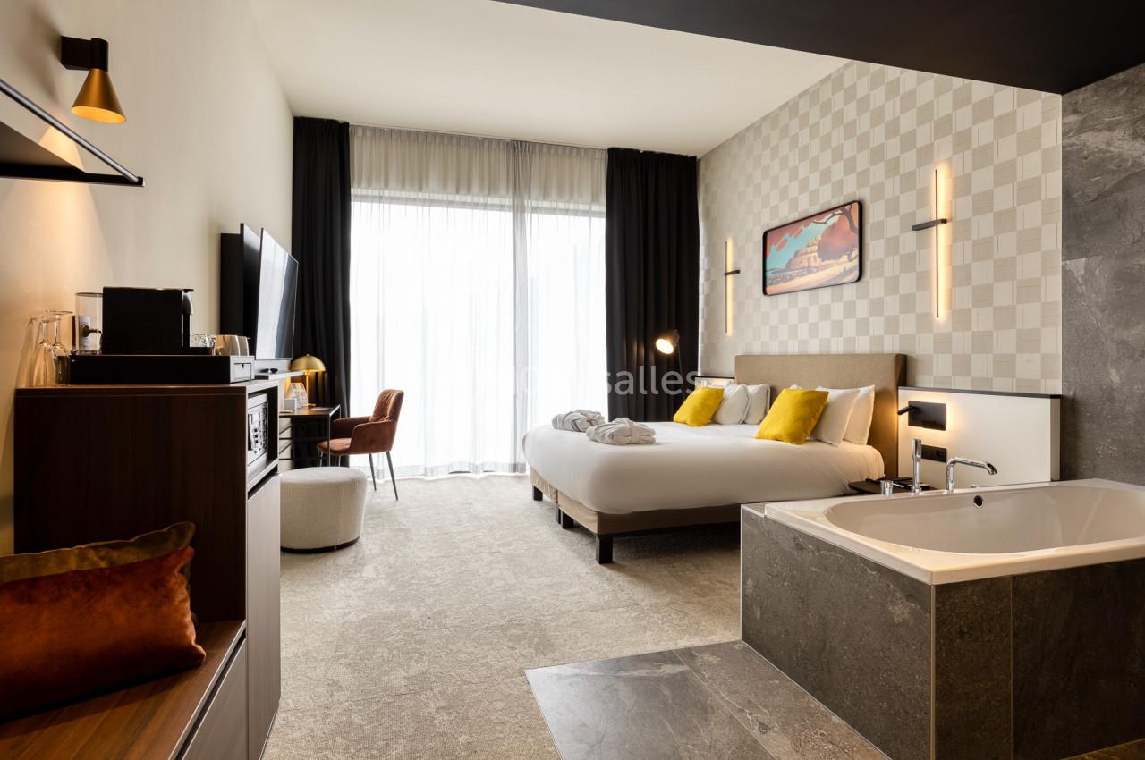 Chambre d'hôtel moderne avec lit double, baignoire intégrée, bureau, télévision et grande baie vitrée.