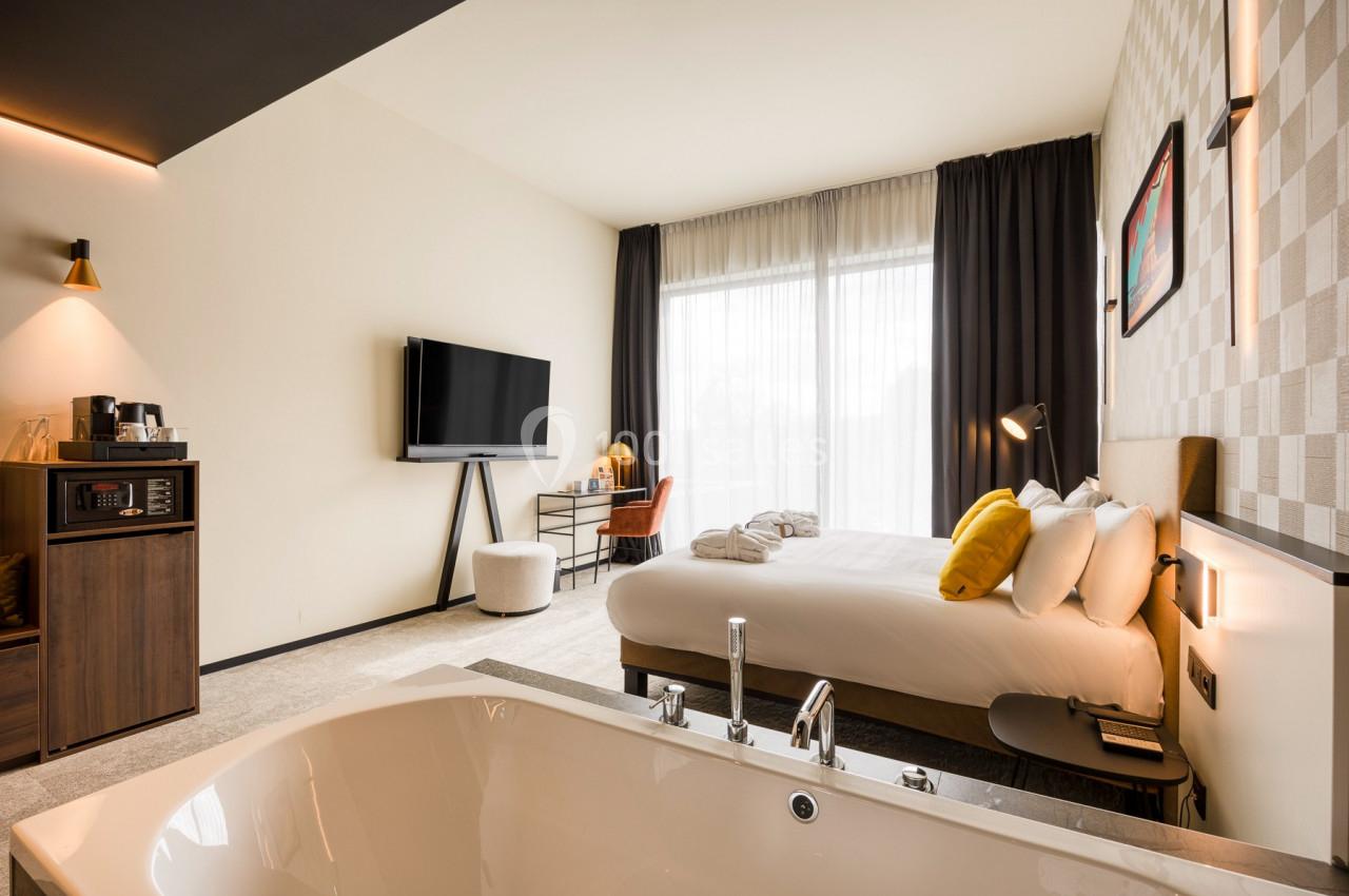 Chambre d'hôtel moderne avec lit double, baignoire au premier plan, bureau, télévision et grande baie vitrée avec rideaux.
