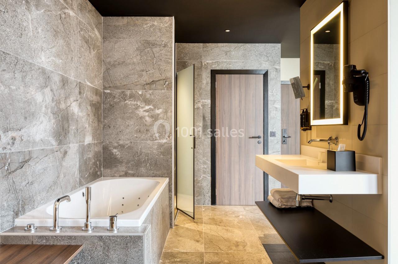 Salle de bain moderne avec baignoire, lavabo suspendu, miroir éclairé et murs en pierre grise.