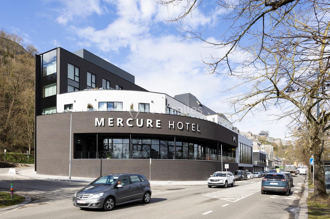 Façade moderne d'un hôtel Mercure avec des voitures circulant sur une route adjacente, entouré d'arbres et de collines.