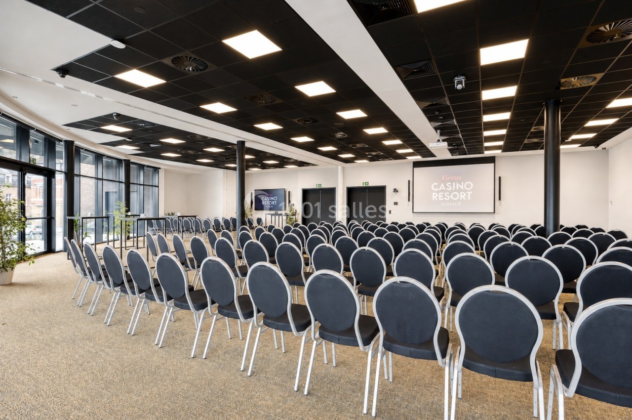 Salle de conférence moderne avec rangées de chaises alignées, écran de projection et grandes baies vitrées.