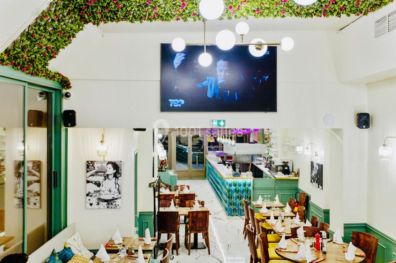 Salle de restaurant lumineuse avec décoration végétale, tables dressées et écran mural diffusant une vidéo.