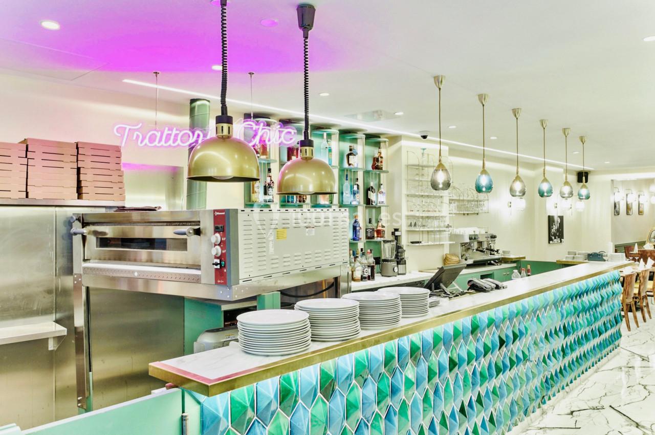 Comptoir de restaurant avec carrelage coloré, four à pizza, étagères garnies et éclairage suspendu.