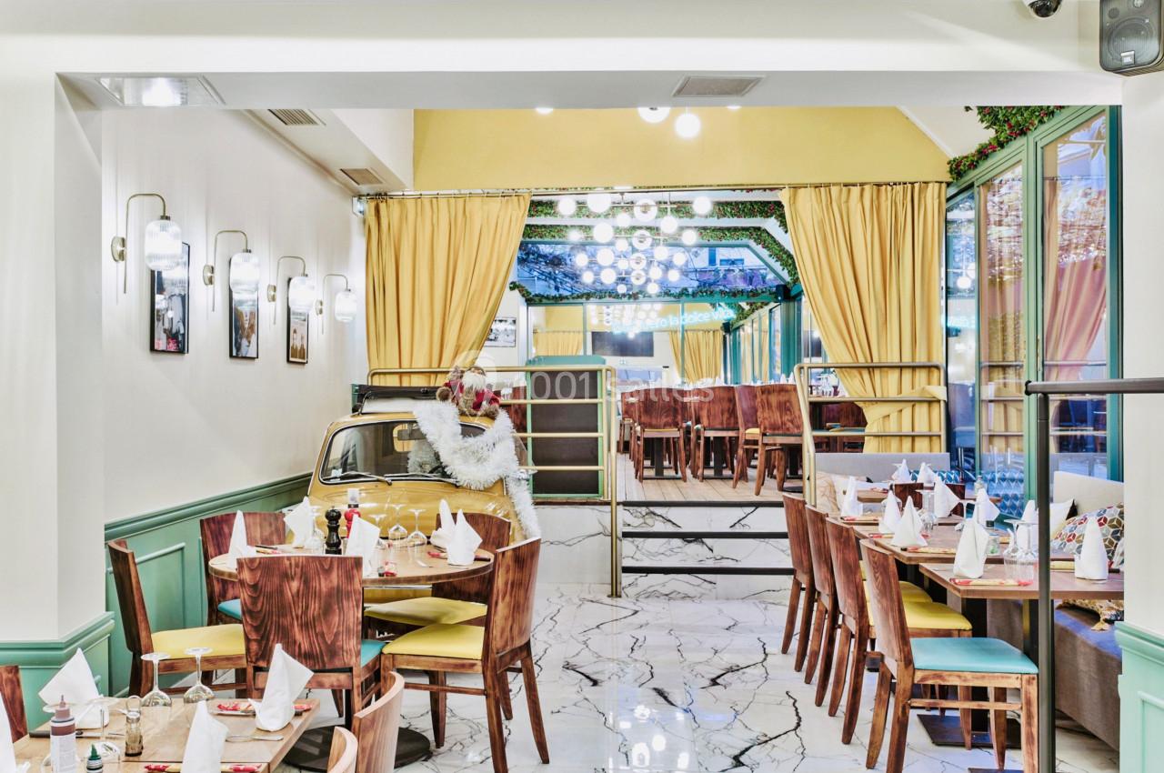 Salle de restaurant lumineuse avec tables dressées, chaises en bois, décorations végétales et rideaux jaunes.