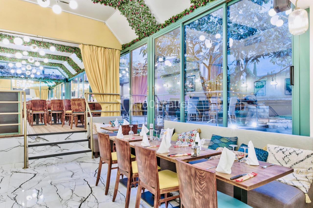 Salle de restaurant lumineuse avec tables dressées, grandes baies vitrées et décoration végétale au plafond.