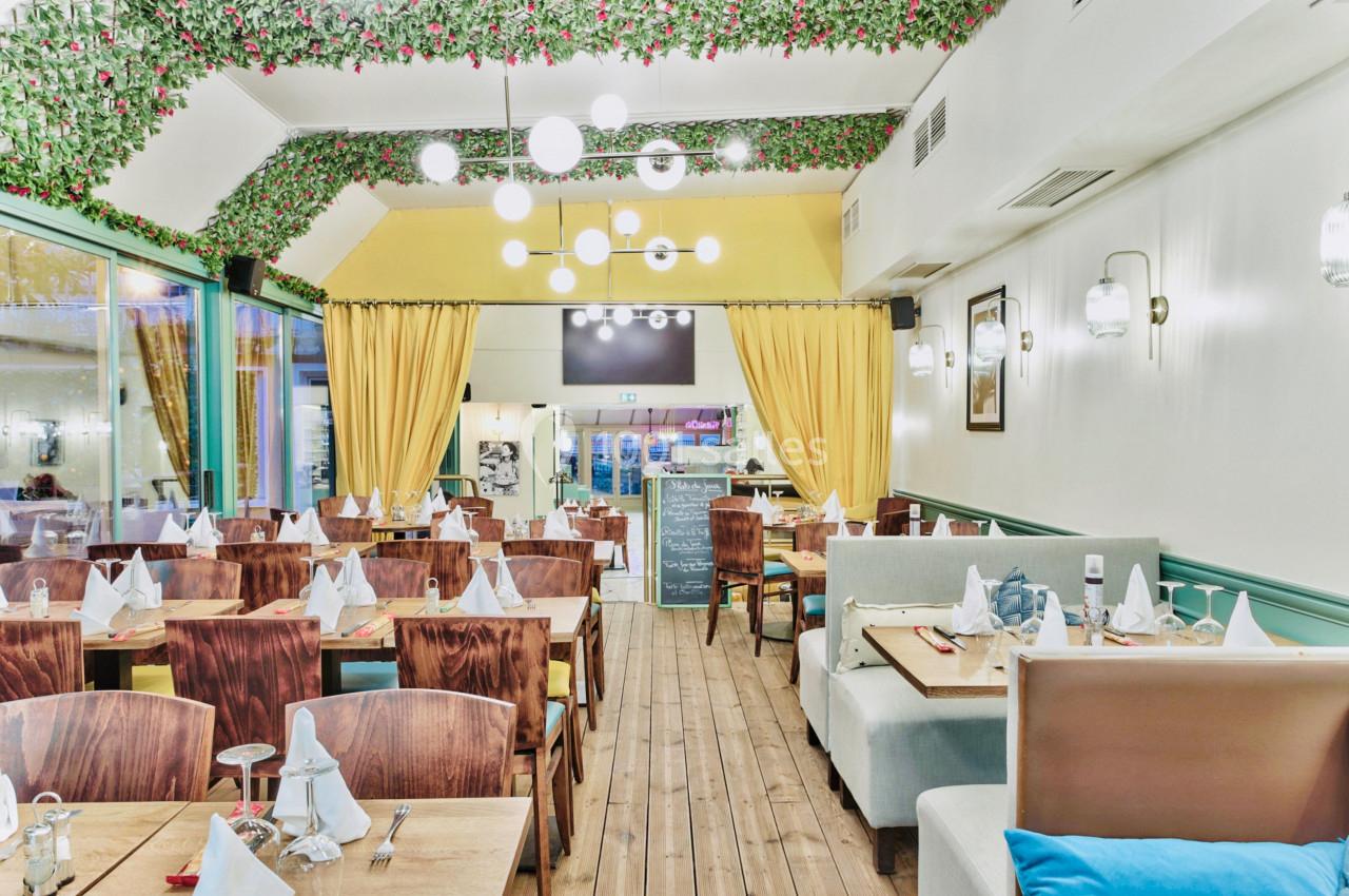 Salle de restaurant lumineuse avec tables dressées, chaises en bois, banquettes et décoration végétale au plafond.