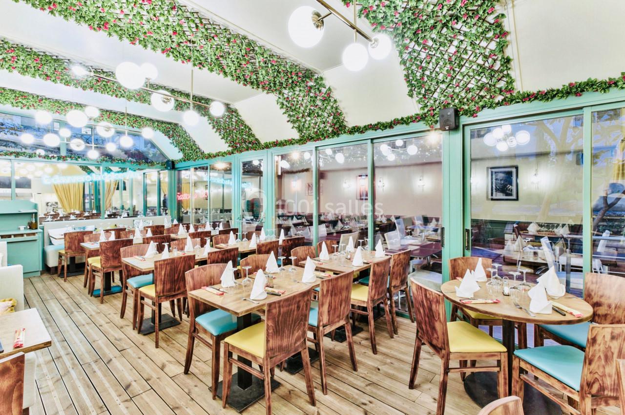 Salle de restaurant lumineuse avec tables en bois dressées, plafond décoré de guirlandes végétales et grandes baies vitrées.