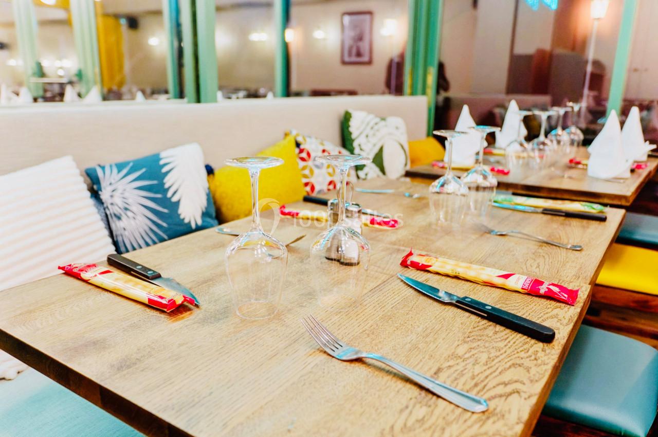Table dressée avec des couverts, verres à pied et serviettes, entourée de coussins colorés dans un restaurant lumineux.