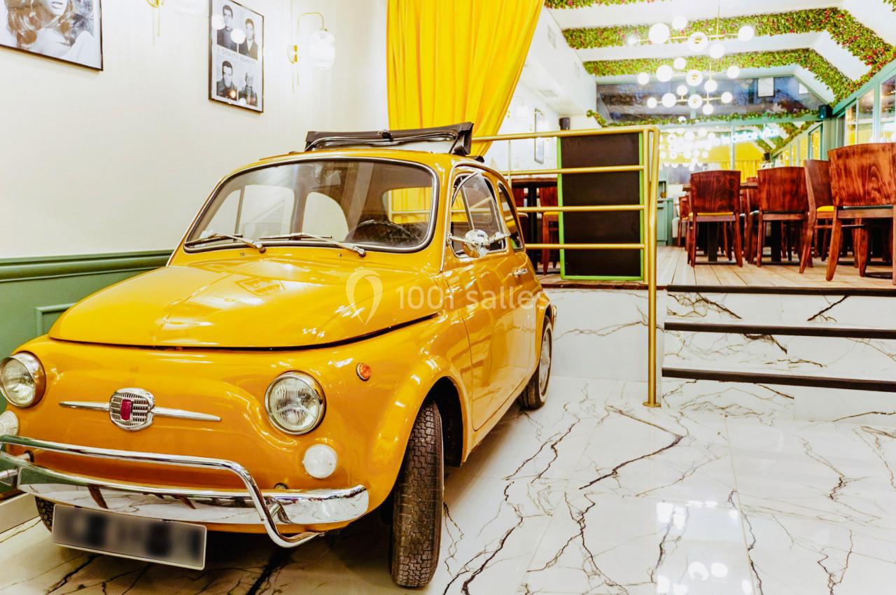 Voiture vintage jaune garée à l'intérieur d'un espace lumineux avec sol en marbre et décoration végétale.