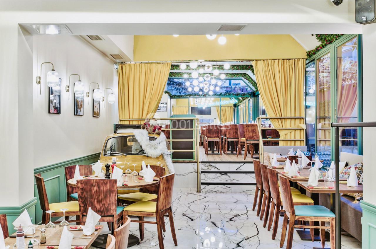 Salle de restaurant lumineuse avec tables dressées, décoration végétale et un espace séparé par des rideaux jaunes.