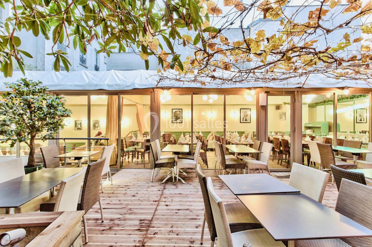 Terrasse en bois aménagée avec des tables et chaises, entourée de végétation et donnant sur une salle lumineuse.
