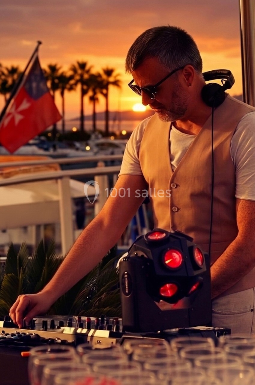 Un DJ mixe en plein air au coucher du soleil, avec des palmiers et un drapeau en arrière-plan.