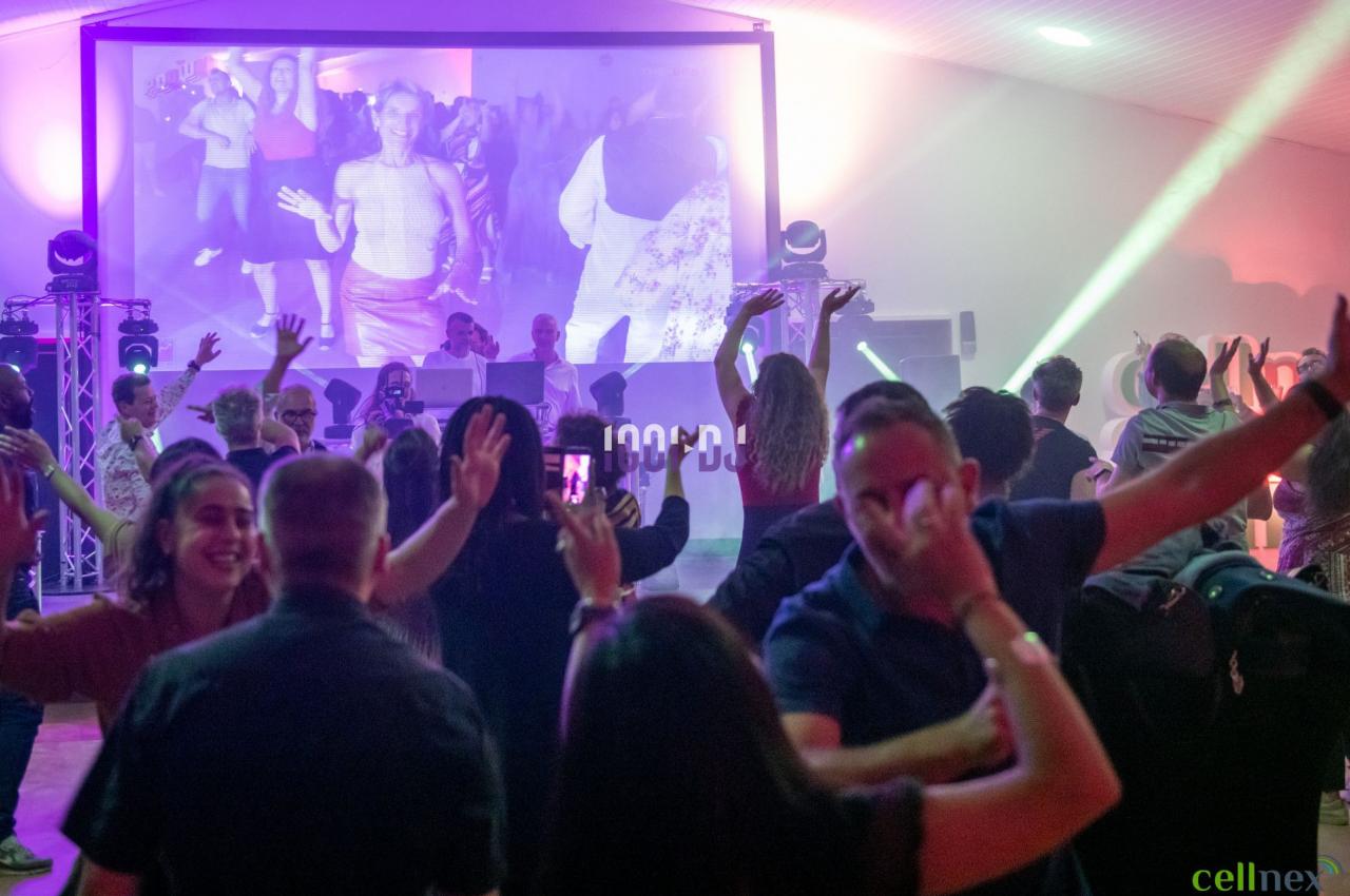 Des personnes dansent dans une salle éclairée par des lumières colorées, avec un écran géant diffusant des images en arrière…