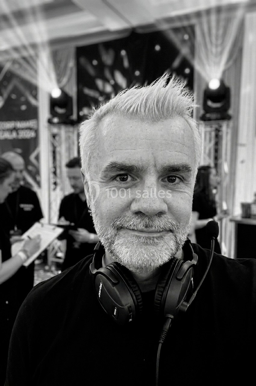 Homme souriant avec un casque audio autour du cou, dans un environnement d'événement avec des lumières et des personnes en…