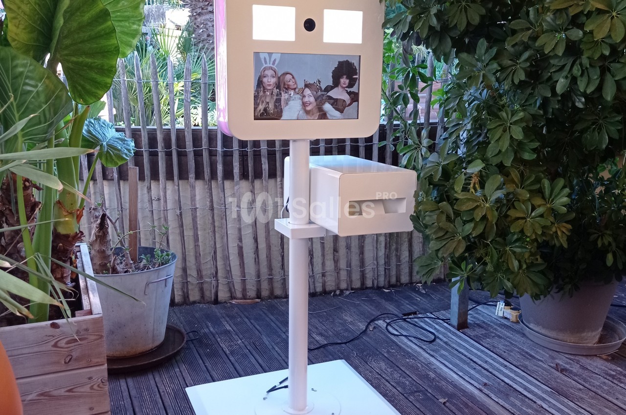 Borne photo interactive sur pied, installée en extérieur sur une terrasse en bois, entourée de plantes.