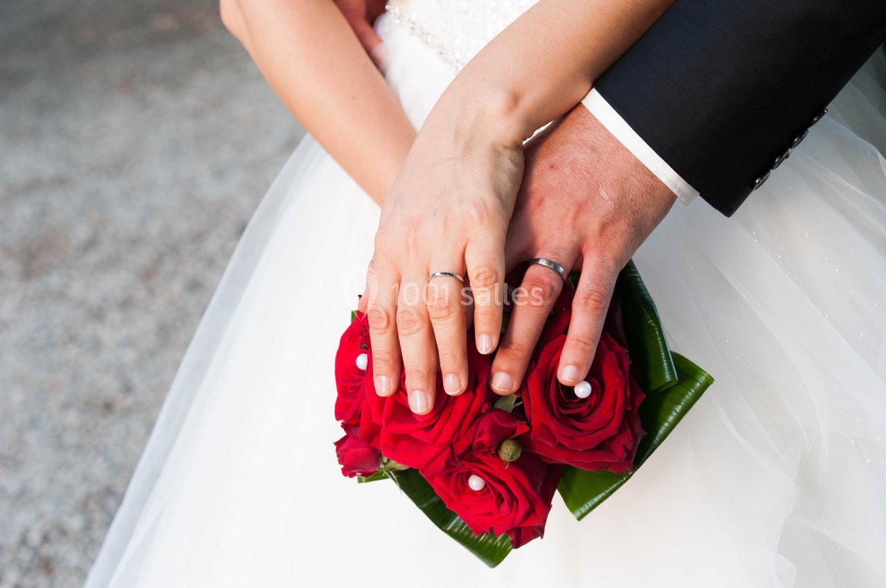 Mains d'un couple marié portant des alliances, posées sur un bouquet de roses rouges.
