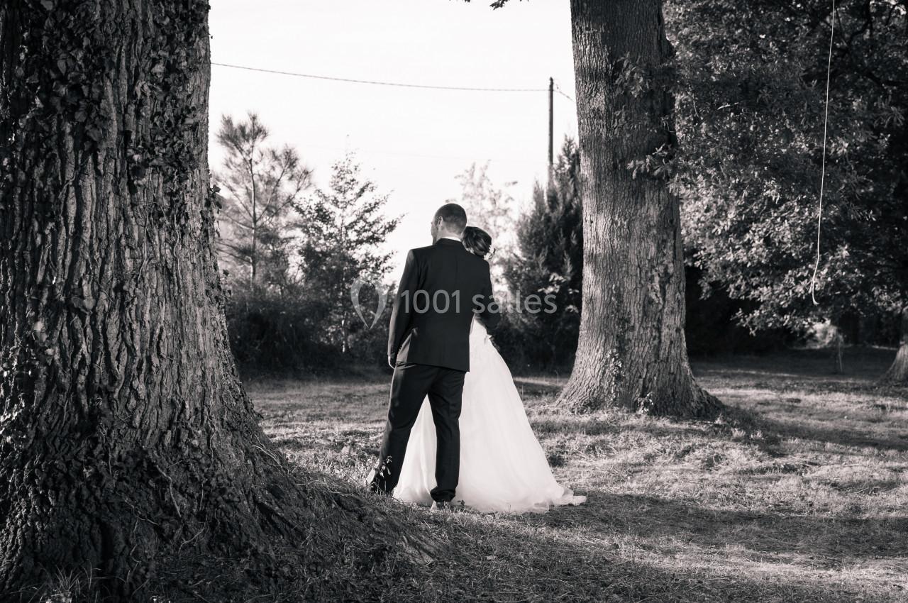Un couple en tenue de mariage marche entre de grands arbres dans un paysage naturel, capturé en noir et blanc.