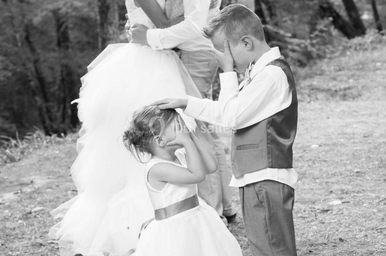 Deux enfants se couvrent les yeux pendant qu'un couple en tenue de mariage s'embrasse dans un cadre boisé.