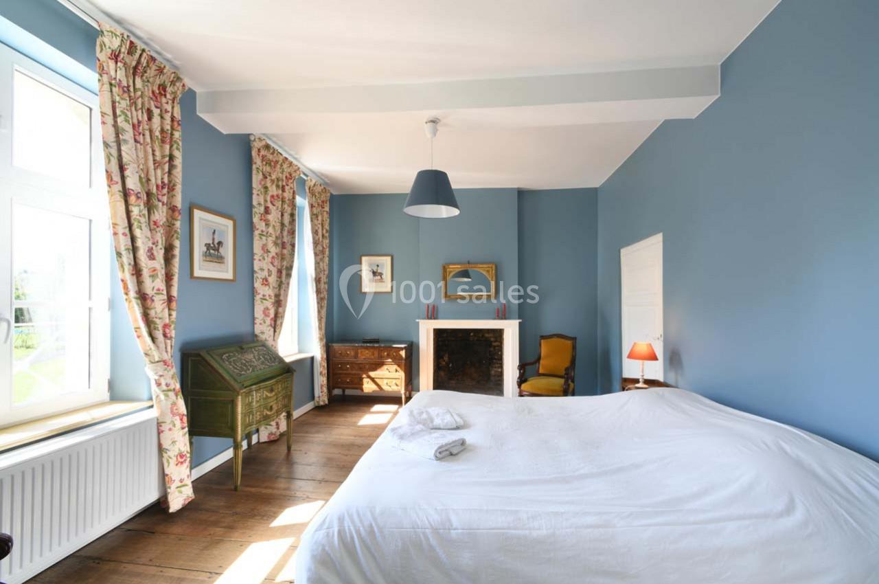 Chambre lumineuse avec murs bleus, lit double, cheminée, bureau en bois, fauteuil jaune et grandes fenêtres.