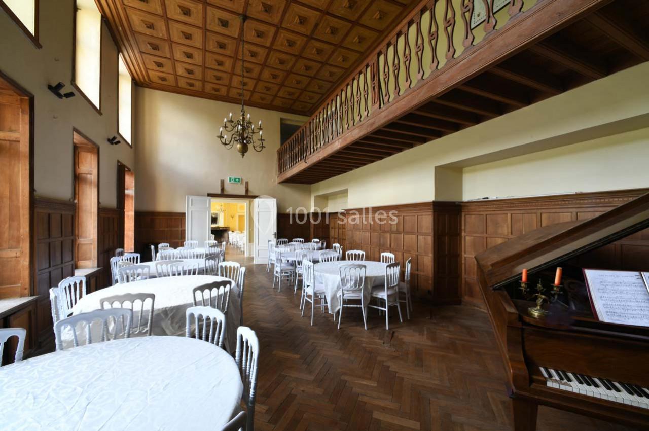 Salle avec parquet en bois, tables rondes dressées, chaises blanches, piano à queue et mezzanine en bois sculpté.