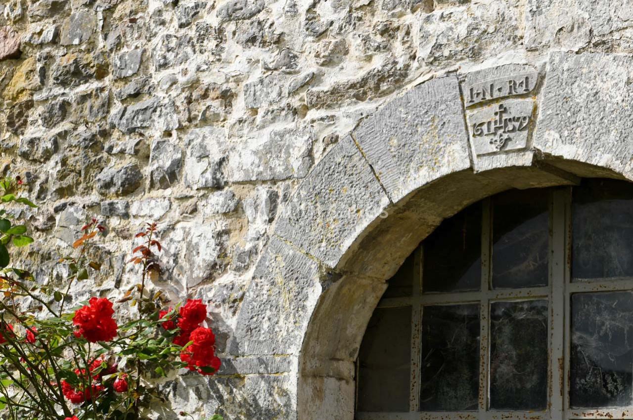 Mur en pierre avec une arche en pierre gravée au-dessus d'une fenêtre, entourée de roses rouges en premier plan.
