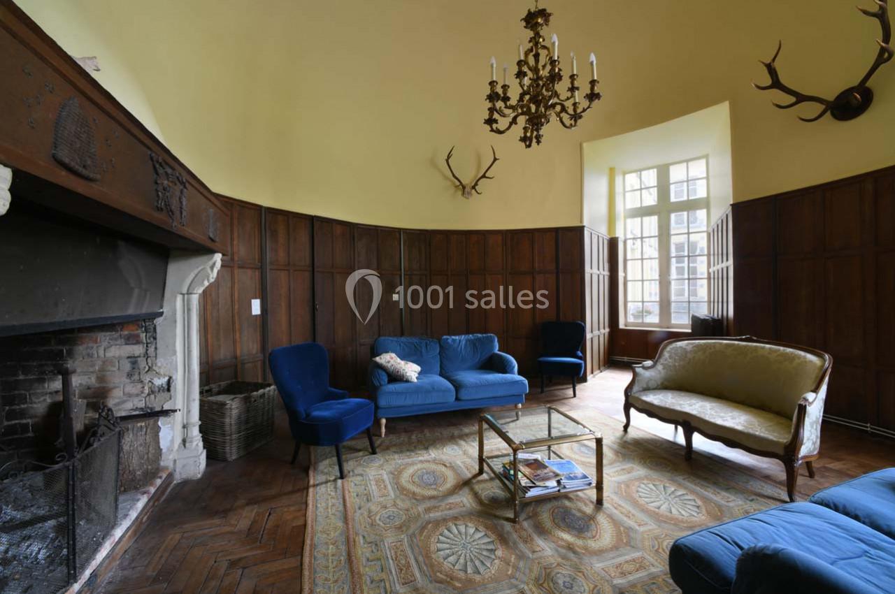 Salon avec murs en bois, cheminée, canapé bleu, fauteuils assortis, lustre, tapis et grande fenêtre lumineuse.
